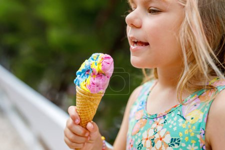 Foto de Feliz niña preescolar comiendo helado colorido en cono de gofre en el día soleado de verano. Pequeño niño pequeño come postre helado. Comida dulce en días calurosos de verano. Luz brillante, helado colorido. - Imagen libre de derechos
