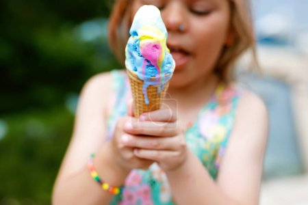 Téléchargez les photos : Bonne fille préscolaire manger de la crème glacée colorée dans un cône de gaufre le jour ensoleillé d'été. Le petit enfant mange du dessert glacé. Nourriture sucrée pendant les chaudes journées d'été. Lumière vive, crème glacée colorée. - en image libre de droit