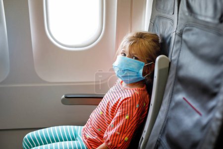 Photo for Little girl with surgical mask face protection traveling by an airplane. Protection against Coronavirus and flu. - Royalty Free Image