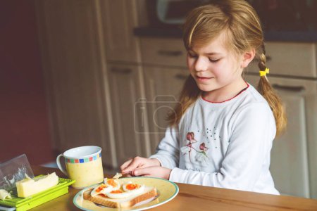 Foto de La niña sonriente desayuna en casa. Niño en edad preescolar comiendo sándwich con huevos cocidos. Niños felices, comida saludable y comida - Imagen libre de derechos
