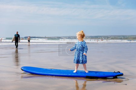 Foto de Pequeña niña linda en la playa de surfistas Ballybunion, divirtiéndose en la tabla de surf por primera vez, costa oeste de Irlanda. Niño feliz disfrutando del verano irlandés con la familia - Imagen libre de derechos
