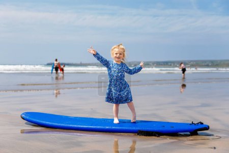 Foto de Pequeña niña linda en la playa de surfistas Ballybunion, divirtiéndose en la tabla de surf por primera vez, costa oeste de Irlanda. Niño feliz disfrutando del verano irlandés con la familia - Imagen libre de derechos