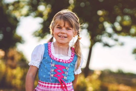 Cute little kid girl in traditional Bavarian costume in wheat field. Happy child with hay bale during Oktoberfest in Munich. Preschool girl play at hay bales during summer harvest time in Germany