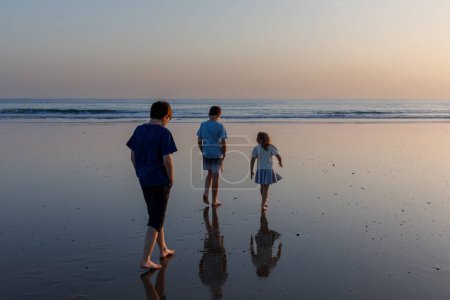 Foto de Tres niños siluetas de pie en la playa al atardecer. familia feliz, dos chicos de la escuela y una niña preescolar. Hermanos divirtiéndose - Imagen libre de derechos