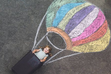 Téléchargez les photos : Joyeux petit enfant volant en montgolfière peint avec des craies colorées aux couleurs arc-en-ciel sur le sol ou de l'asphalte en été. Mignon enfant qui s'amuse. loisirs créatifs pour les enfants - en image libre de droit