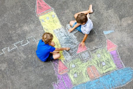 Téléchargez les photos : Deux petits enfants dessinent un château de chevalier avec des craies colorées sur asphalte. Heureux frères et sœurs et amis qui s'amusent à créer une image et une peinture à la craie. Loisirs créatifs pour les enfants en été - en image libre de droit