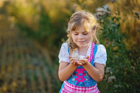 Photo for Cute little kid girl in traditional Bavarian costume in wheat field. Happy child with hay bale during Oktoberfest in Munich. Preschool girl play at hay bales during summer harvest time in Germany - Royalty Free Image