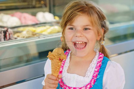 Foto de Little preschool girl eating sweet ice cream in waffle cone on sunny summer day. Happy toddler child eat icecream dessert. Sweet food on hot warm summertime days. - Imagen libre de derechos