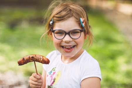 Photo for Little preschool girl eating grilled sausage. Happy child on barbecue or picknick. Healthy food, family summer time - Royalty Free Image