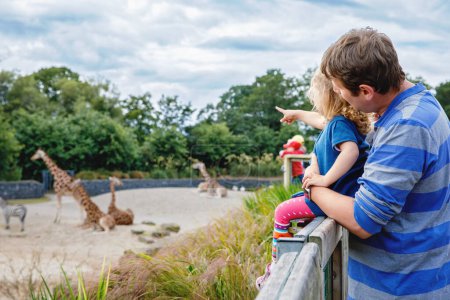 Téléchargez les photos : Mignon adorable tout-petit fille et père regarder et nourrir girafe dans zoo. Bébé enfant heureux, fille et papa, famille s'amuser avec les animaux safari parc sur chaude journée d'été. Irlande. - en image libre de droit