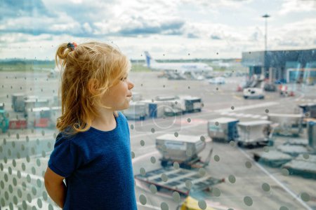 Téléchargez les photos : Jolie petite fille tout-petit à l'aéroport, voyageant. Heureux enfant en bonne santé attendant près de la fenêtre et regardant les avions. Famille en vacances d'été en avion - en image libre de droit