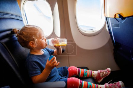 Téléchargez les photos : Adorable petite fille voyageant en avion. Petit enfant en bas âge buvant du jus d'orange assis près de la fenêtre de l'avion. Voyager à l'étranger avec des enfants. Famille en vacances d'été - en image libre de droit