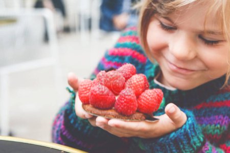 Foto de Adorable niña y delicioso pastel de frambuesa. Niños en edad preescolar comiendo postre dulce en la cafetería francesa al aire libre - Imagen libre de derechos