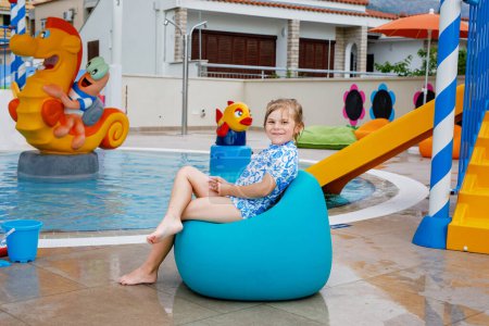 Téléchargez les photos : Petite fille d'âge préscolaire assise près de la piscine extérieure dans la station hôtelière. Enfant apprendre à nager dans la piscine extérieure, éclabousser d'eau, profiter, rire et s'amuser. Vacances en famille - en image libre de droit