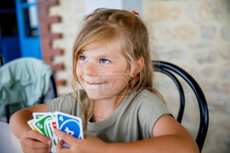 Foto de Emocionado sonriente linda niña preescolar jugando juego de cartas. Feliz niño sano juega con la familia. Creativo en interiores de ocio y educación de los niños. Actividad familiar en casa - Imagen libre de derechos
