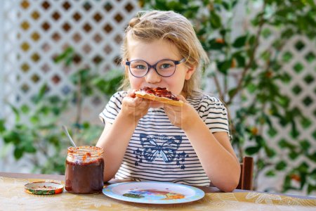 Photo for Cute funny preschool girl eats sweet bun for breakfast. Happy child eating bread roll with strawberry, cherry or plum jam. Health food for children and kids with selfmade jelly. - Royalty Free Image