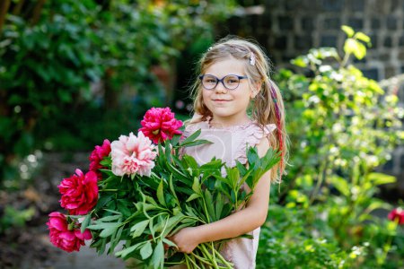Téléchargez les photos : Jolie adorable petite fille préscolaire avec un énorme bouquet de fleurs de pivoine rouge et rose en fleurs. Portrait d'un enfant souriant d'âge préscolaire dans un jardin domestique le jour chaud du printemps ou de l'été. L'été - en image libre de droit