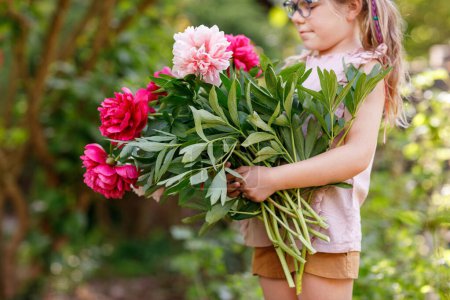 Photo for Cute adorable little preschool girl with huge bouquet of blossoming red and pink peony flowers. Portrait of smiling preschool child in domestic garden on warm spring or summer day. Summertime - Royalty Free Image