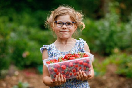 Photo for Happy preschool girl holding box with healthy strawberries from organic berry farm in summer, on sunny day. Smiling child. Kid with fresh ripe red berries, eats strawberry - Royalty Free Image
