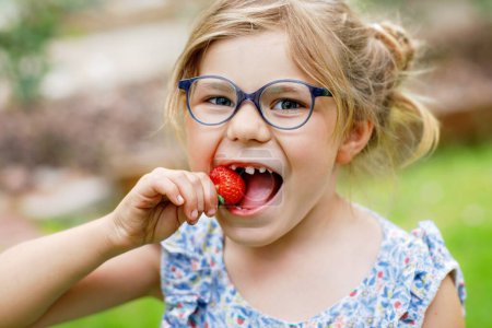 Photo for Happy preschool girl eating fresh strawberry. Smiling child with glasses biting in a ripe red berry. Healthy food and children - Royalty Free Image