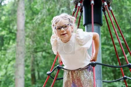 Photo for Adorable happy little girl is climbing on an outdoor playground on a summer day. Blonde preschool child climbs to the top of the game complex. - Royalty Free Image