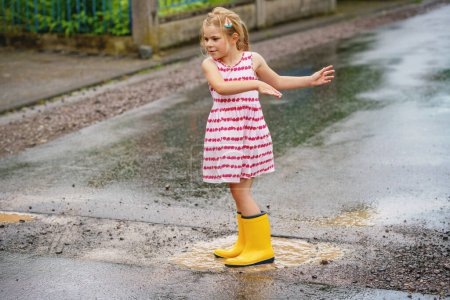 Téléchargez les photos : Joyeux petit enfant d'âge préscolaire portant des bottes de pluie jaunes et marchant pendant les flaques d'eau. Enfant mignon en vêtements colorés sautant dans la flaque d'eau, éclaboussures d'eau, activité de plein air. - en image libre de droit