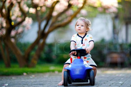 Foto de Linda niña jugando con azul pequeño coche de juguete en el jardín de casa o guardería. Adorable niño hermoso niño con magnolia floreciente en el fondo. Juego activo al aire libre con niños - Imagen libre de derechos
