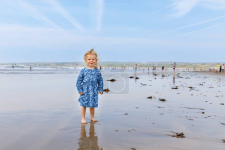 Foto de Little cute toddler girl at the Ballybunion surfer beach, having fun on with playing on west coast of Ireland. Happy child enjoying Irish summer and sunny day with family - Imagen libre de derechos