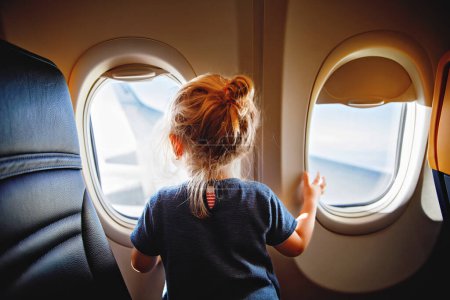Adorable little girl traveling by an airplane. Child sitting by aircraft window and looking outside. Traveling with kids abroad. Family on summer vacations