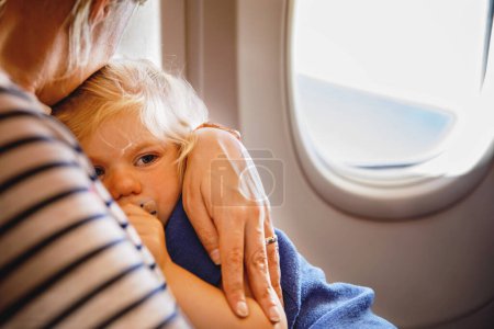 Téléchargez les photos : Femme voyageant avec un petit enfant en avion. Triste petite fille fatiguée assise avec maman par la fenêtre de l'avion. Concept de maternité. Pleurer bébé - en image libre de droit