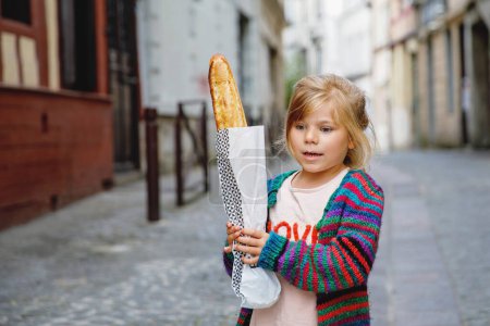 Foto de Adorable niña preescolar con baguette francesa fresca en el lado de la calle de la ciudad. Feliz niño pequeño en París, Francia - Imagen libre de derechos