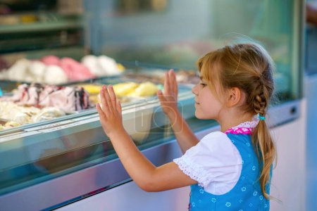 Téléchargez les photos : Mignon petite fille préscolaire choisir et acheter de la crème glacée dans le café stand extérieur. Joyeux enfant souriant regardant différentes sortes de glace. Doux dessert d'été. - en image libre de droit