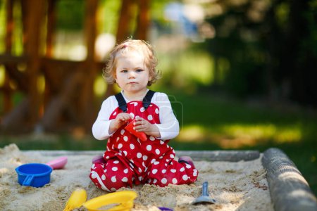 Téléchargez les photos : Petite fille mignonne jouant dans le sable sur une aire de jeux extérieure. Beau bébé en chewing-gum rouge qui s'amuse par une chaude journée d'été ensoleillée. Enfant avec des jouets de sable colorés. Bébé actif sain à l'extérieur joue à des jeux - en image libre de droit
