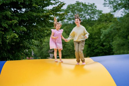 Téléchargez les photos : Petite fille d'âge préscolaire et sœur d'école sautant sur le trampoline. Heureux enfants drôles, frères et sœurs amoureux s'amuser avec l'activité de plein air en été. Trampoline aux couleurs du drapeau ukrainien. - en image libre de droit