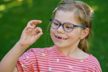Téléchargez les photos : Portrait d'une jolie fille d'âge préscolaire avec des lunettes et de grandes dents à l'extérieur dans le parc. Joyeux enfant drôle sur une journée ensoleillée d'été. Enfant perdant une dent de lait. - en image libre de droit