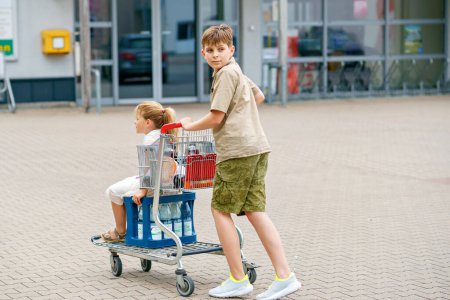 Photo for A School Boy Pushes the Cart While a Preschool Girl Happily Sits Inside, as They Enjoy a Fun Family Shopping Trip. Happy Siblings Outdoors, in Front of a Supermarket. - Royalty Free Image