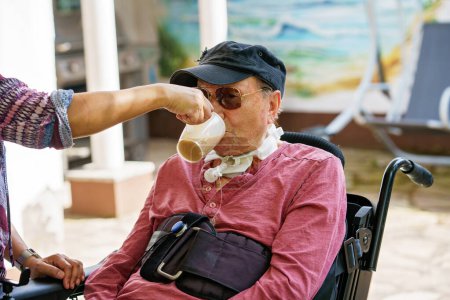 Photo for Senior man of 85 years in an electric wheelchair is sitting in a park. Nurse supporting man with drinking coffee, holding cup - Royalty Free Image