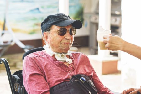 Téléchargez les photos : Un aîné de 85 ans en fauteuil roulant électrique est assis dans un parc. Infirmière soutenant l'homme avec boire du café, tenant tasse - en image libre de droit