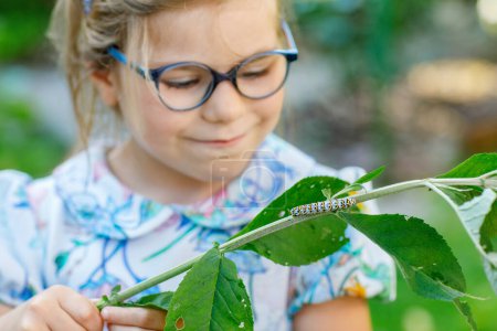 Foto de Pequeña niña preescolar observa oruga subir a la planta. Feliz niño emocionado viendo y aprendiendo insectos en jardines domésticos - Imagen libre de derechos