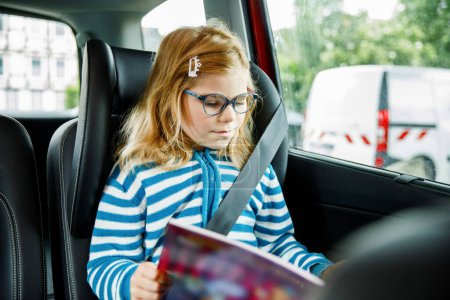 Photo for Little Preschool Girl Sitting in Her Car Seat. Happy Child with Eyeglasses Reading a Book, Smiling on the Way to Family Vacations during Traffic Jam - Royalty Free Image