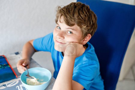 Téléchargez les photos : Joyeux garçon blond préadolescent qui mange des céréales pour le petit déjeuner ou le déjeuner. Manger sainement pour les enfants le matin. Ecole enfant petit déjeuner avec lait et avoine granola muesli. - en image libre de droit
