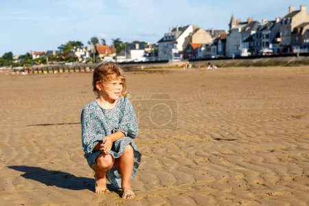 Foto de Preschool Girl Plays on a Norman Beach in Normandy, Embracing the Joy of Sandy Shores, Seashells, and Carefree Exploration (en inglés). Felices vacaciones de niños y familias en Francia - Imagen libre de derechos