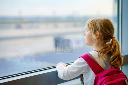 Téléchargez les photos : Petite fille à l'aéroport attendant l'embarquement à la grande fenêtre. Cute Kid se tient à la fenêtre dans le contexte des avions. Nous avons hâte de partir pour des vacances d'été en famille. - en image libre de droit