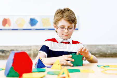 Foto de Niño pequeño con gafas jugando con elementos plásticos coloridos kit en la escuela o guardería preescolar. Feliz niño construyendo y creando figuras geométricas, aprendiendo matemáticas y geometría - Imagen libre de derechos