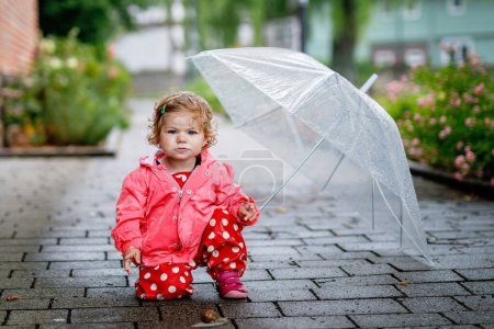 Foto de Linda niña adorable descubrir caracol en un paseo. Hermoso niño rizado que se divierte en el día lluvioso. Con paraguas grande, niño en ropa impermeable
. - Imagen libre de derechos