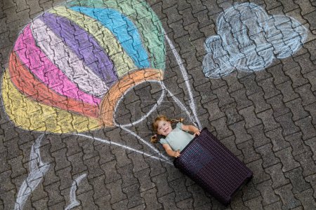 Foto de Pequeña niña feliz volando en globo aerostático pintado con tiza de colores en colores arcoíris en el suelo o asfalto en verano. Lindo niño divirtiéndose. ocio creativo para niños - Imagen libre de derechos
