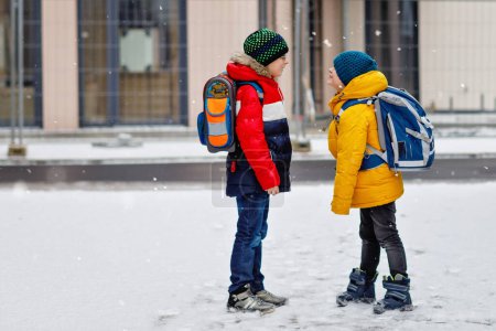 Photo for Two little kids boys of elementary class walking to school during snowfall. Happy children having fun and playing with first snow. Siblings and best friends with backpack in colorful winter clothes - Royalty Free Image