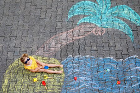 Téléchargez les photos : Happy little preschool girl in swimsuit with sea, sand, palm painted with colorful chalks on asphalt. Cute child with having fun with chalk picture. Summer, vacations, summertime. - en image libre de droit