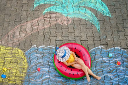 Foto de Happy little preschool girl in swimsuit on inflatable ring with sea, sand, palm painted with colorful chalks on asphalt. Cute child with having fun with chalk picture. Summer, vacations, summertime. - Imagen libre de derechos