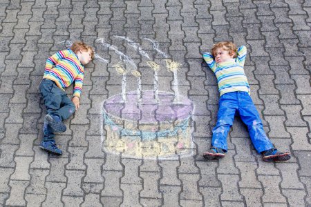 Foto de Dos niños pequeños felices que se divierten con el dibujo grande de la imagen del pastel de cumpleaños con tiza de colores. Ocio creativo para los niños al aire libre en verano. Niños soplando velas
. - Imagen libre de derechos
